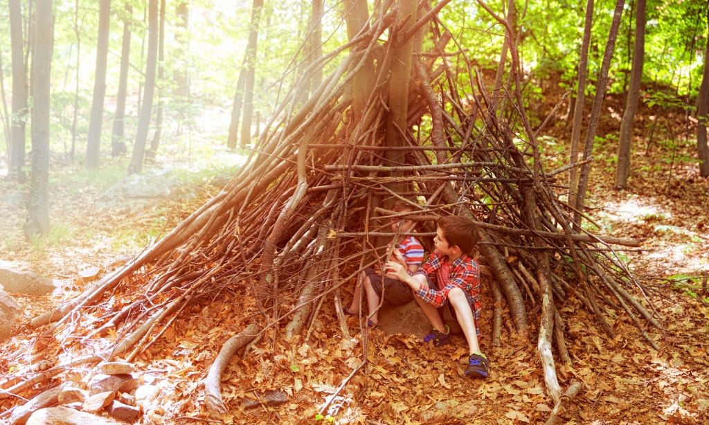 Zwei Kinder haben in einem Laubwald eine Hütte aus Ästen gebaut. 