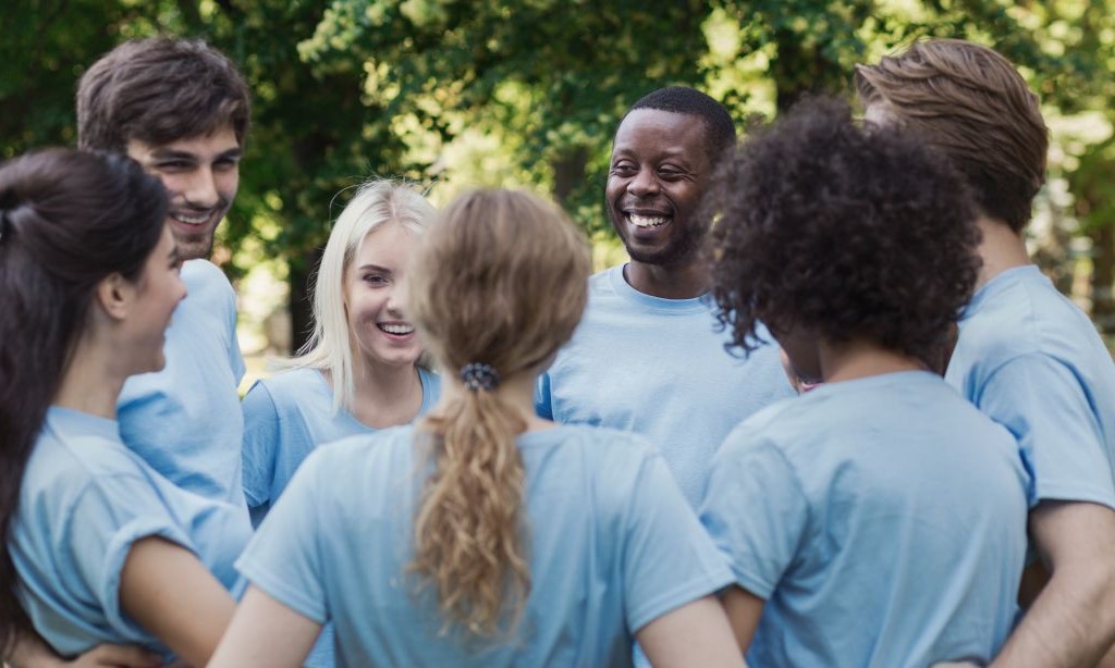 Sieben Personen in blauen T-Shirts stehen im Kreis und haben die Arme umeinander gelegt, während sie reden. 