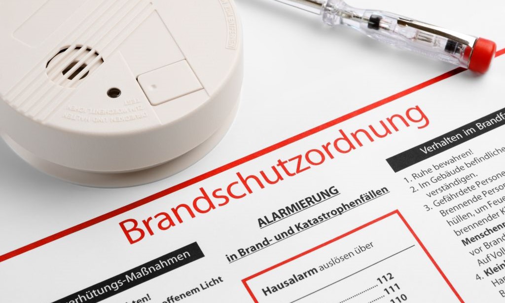 Auf einem weißen Tisch liegen eine Brandschutzordnung, ein Schraubenzieher und ein Feuermelder. 