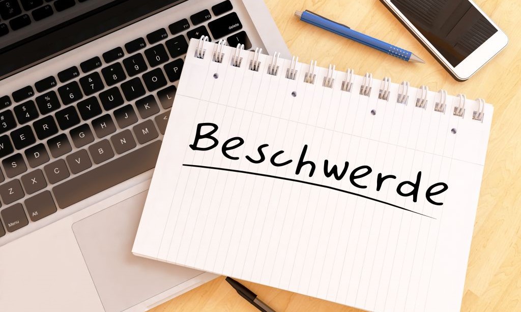 Auf einem Notizblock steht „Beschwerde“. Der Block liegt auf einem Laptop und daneben liegen ein Kugelschreiber und ein Handy.