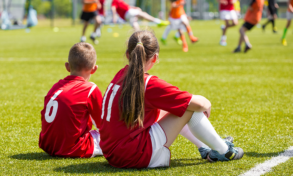 Zwei Kinder in Fußballkleidung schauen anderen Kindern beim Fußballspielen zu. 