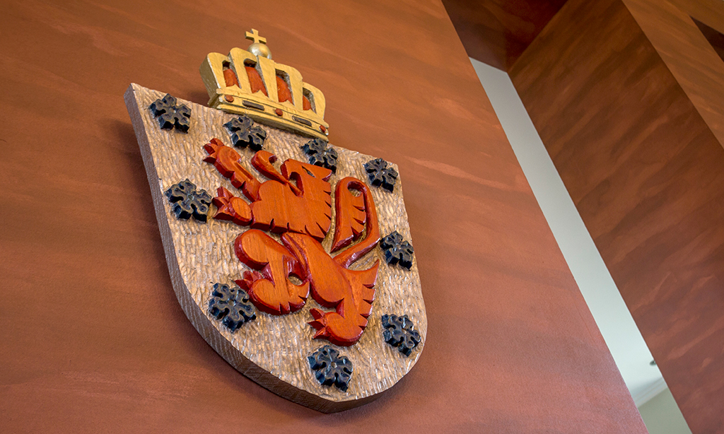Wappen der Deutschsprachigen Gemeinschaft mit seinem roten Löwen umrandet von blauen Blüten und oberhalb abgeschlossen mit einer goldenen Krone hängt an einer Wand. 