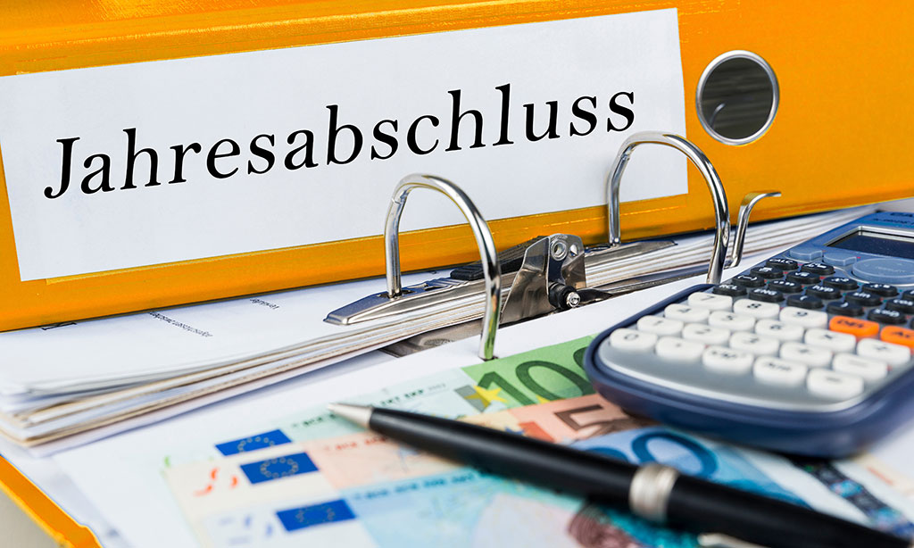 Ein orangener Ordner mit der Aufschrift „Jahresabschluss“, ein Taschenrechner, ein Kugelschreiber und drei Gelscheine liegen auf einem geöffneten Ordner. 