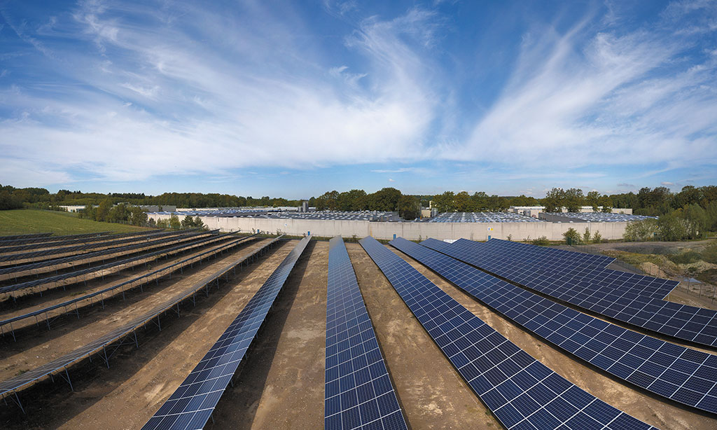 Ein Solarpark steht auf einer Wiese, im Hintergrund ist ein Fabrik zu sehen.