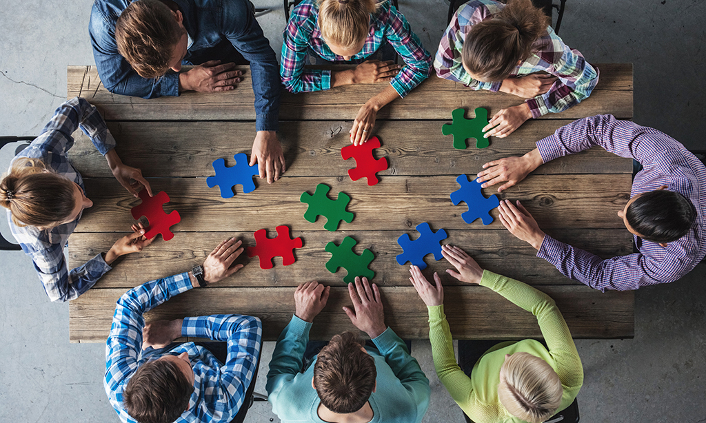 Eine Gruppe von Menschen sitzt an einem Tisch. Von oben sieht man, wie sie gemeinsam an einem Puzzle sitzen.