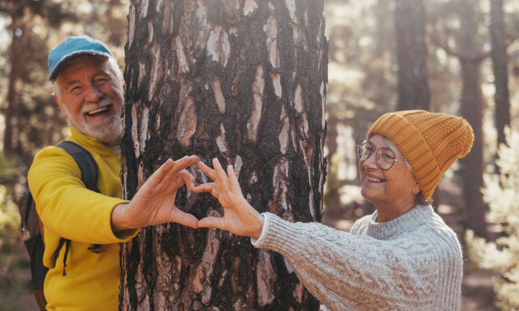 Ein älterer Mann und eine ältere Frau stehen neben einem Baum und halten ihre Hände so zusammen, dass sie eine Herzform ergeben.