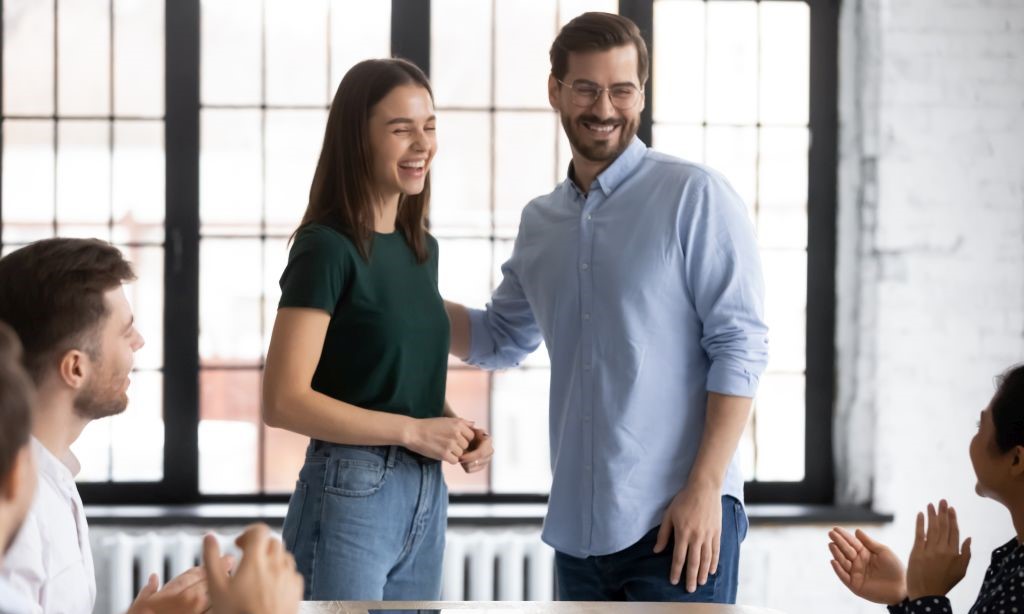 Ein Mann und ein junges Mädchen stehen vor einem Versammlungstisch, an dem Personen sitzen und klatschen. Der Mann hat seinen Arm auf den Arm des Mädchens gelegt und beide lachen.