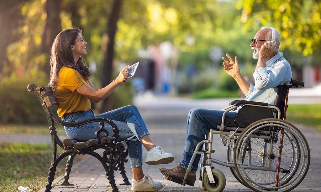 Eine Frau sitzt auf eine Parkbank und bedient ein Tablet, während ihr ein Mann im Rollstuhl mit Kopfhörern gegenüber sitzt.