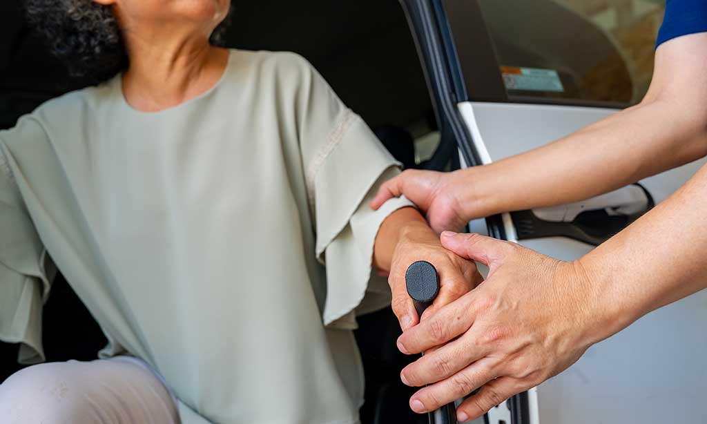 Eine Frau mit Stock steigt mit Hilfe aus einem Auto aus