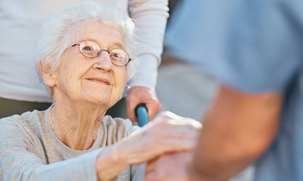 Eine alte Frau im Rollstuhl hält eine Hand und schaut zu einer anderen Person hoch.