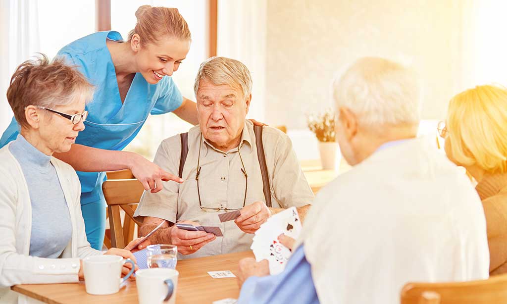 Eine Pflegekraft steht am Tisch mit älteren Menschen, die Karten spielen.