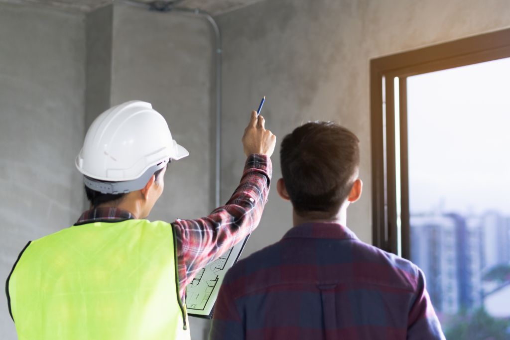 Zwei Männer begutachten eine Wand mit einem Fenster. Ein Mann mit Leuchtweste und Helm zeigt auf die Wand.