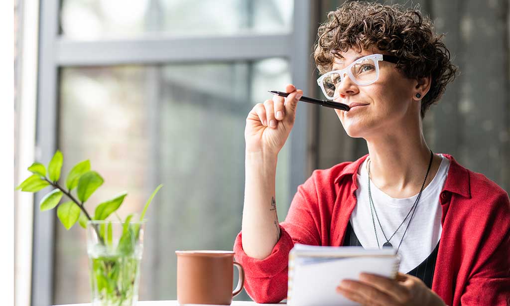 Eine Frau mit Brille sitzt an einem Tisch und hält in der einen Hand einen Stift, den Sie an ihren Mund anlehnt, und in der anderen ein Notizheft. 