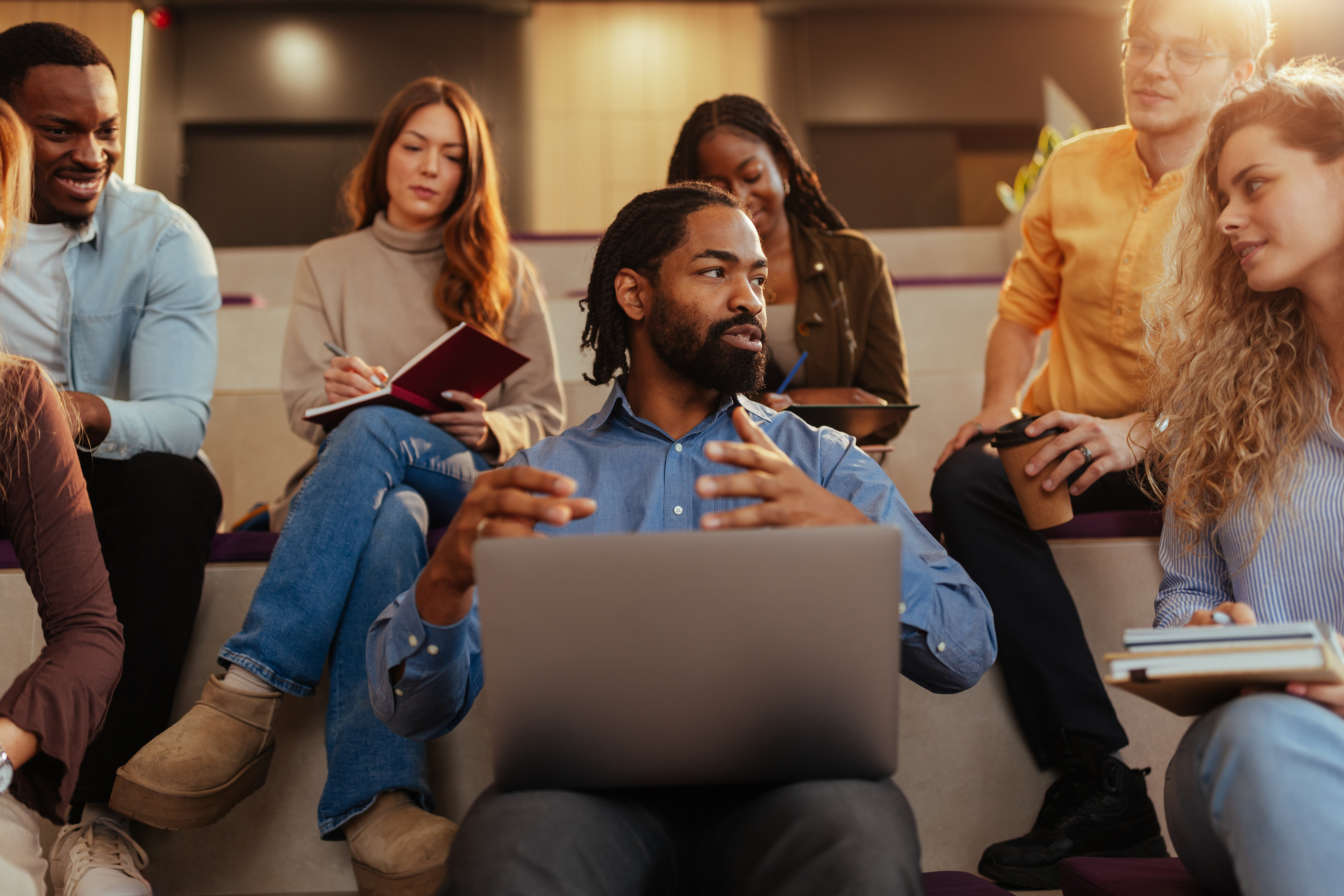 Ein Mann sitzt mit aufgeklapptem Laptop auf den Knien und gestikuliert mit den Händen zu einer Frau hingewandt, die rechts von im sitzt. Hinter beiden Personen, sitzen noch weitere Personen. 