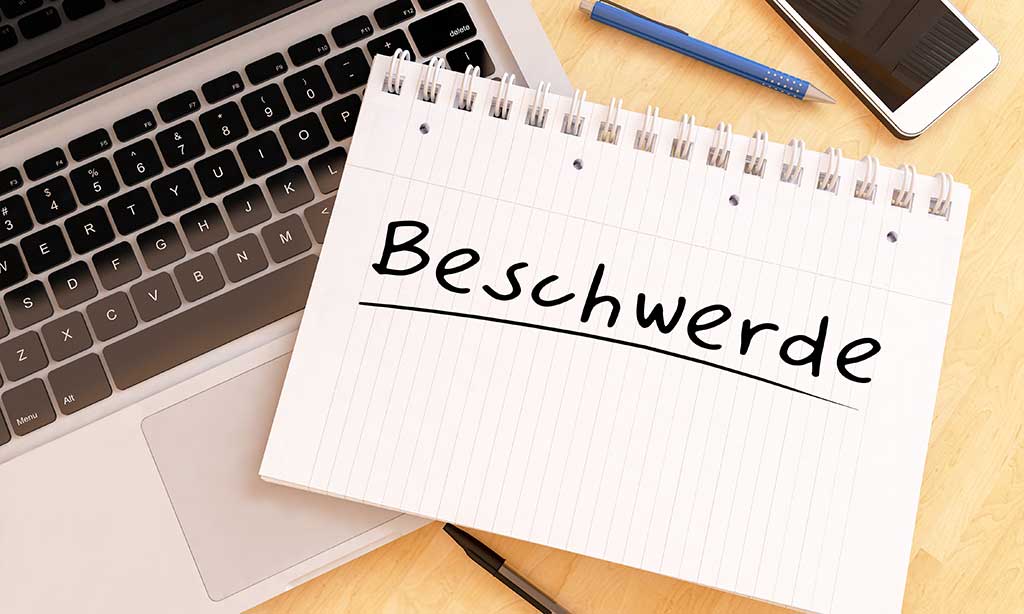 Vogelperspektive auf einen Tisch, auf dem ein aufgeklappter Laptop steht, auf dem ein Schreibblock mit der Aufschrift „Beschwerde“ liegt. 