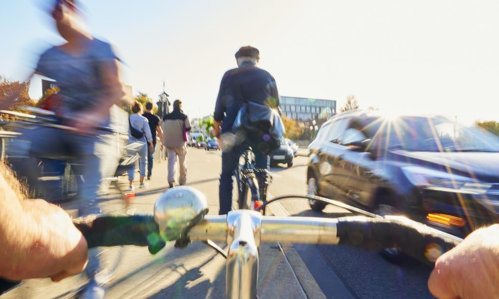Man sieht Autos auf einer Straße und Fußgänger und Fahrradfahrer auf dem Bürgersteig.