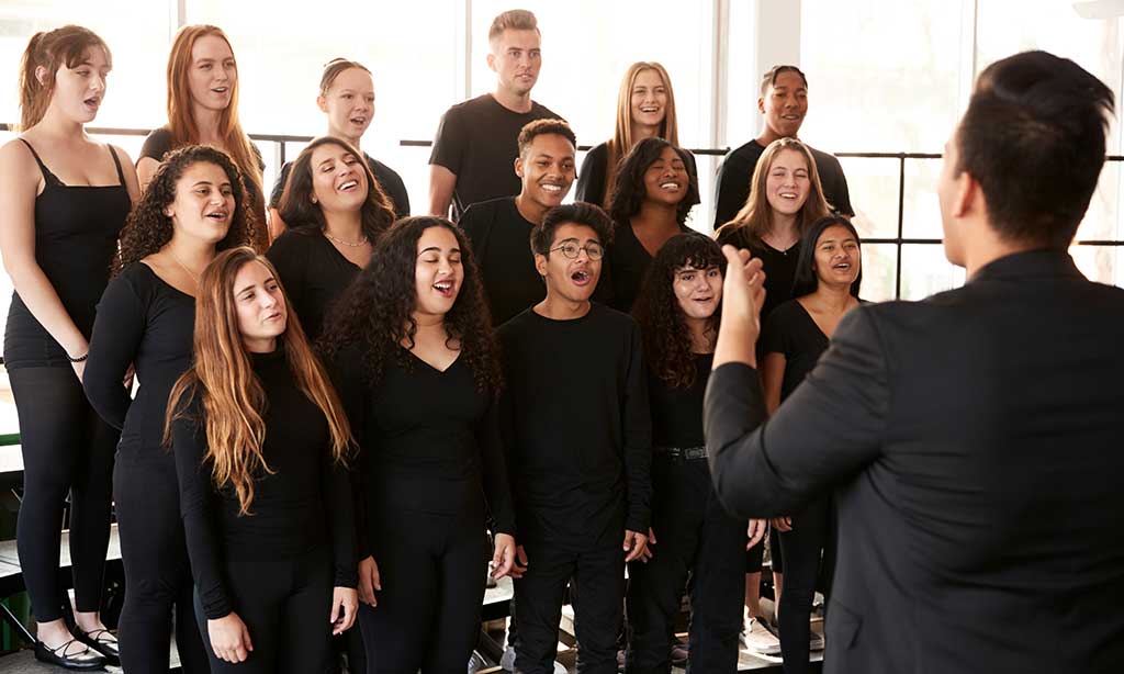 Männer und Frauen singen in einem Chor und werden von einem Dirigenten angeleitet. 