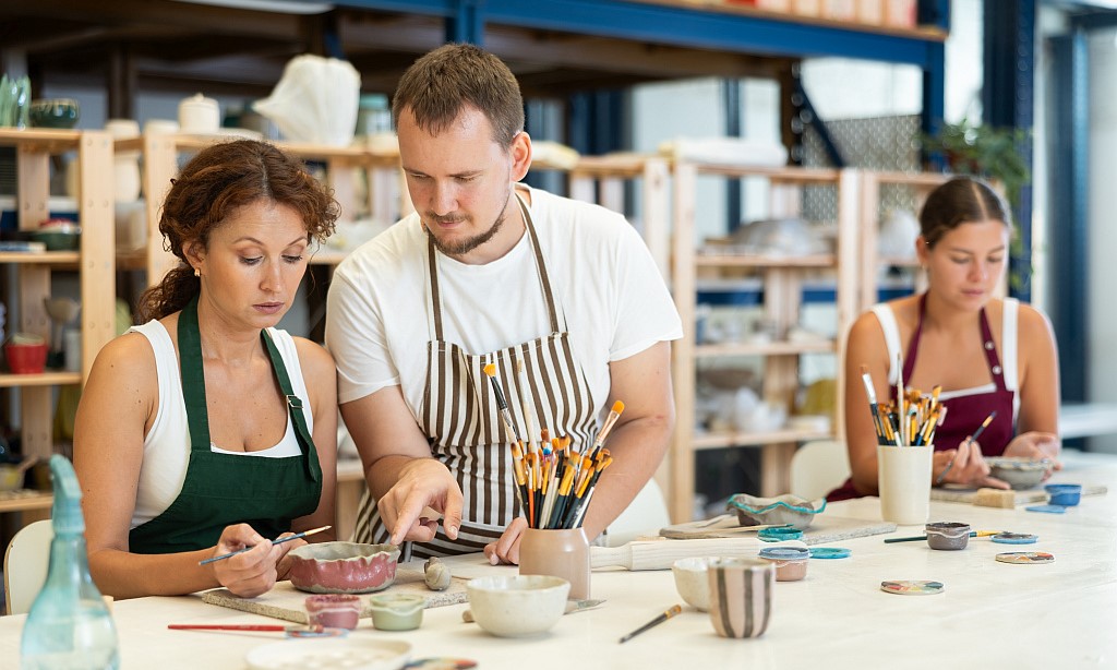 Zwei Frauen nehmen an einem Töpferkurs teil. Sie sitzen an einem Tisch, vor ihnen stehen Tongefäße und Arbeitsmaterial. Ein Mann gibt einer der Frauen Erklärungen.