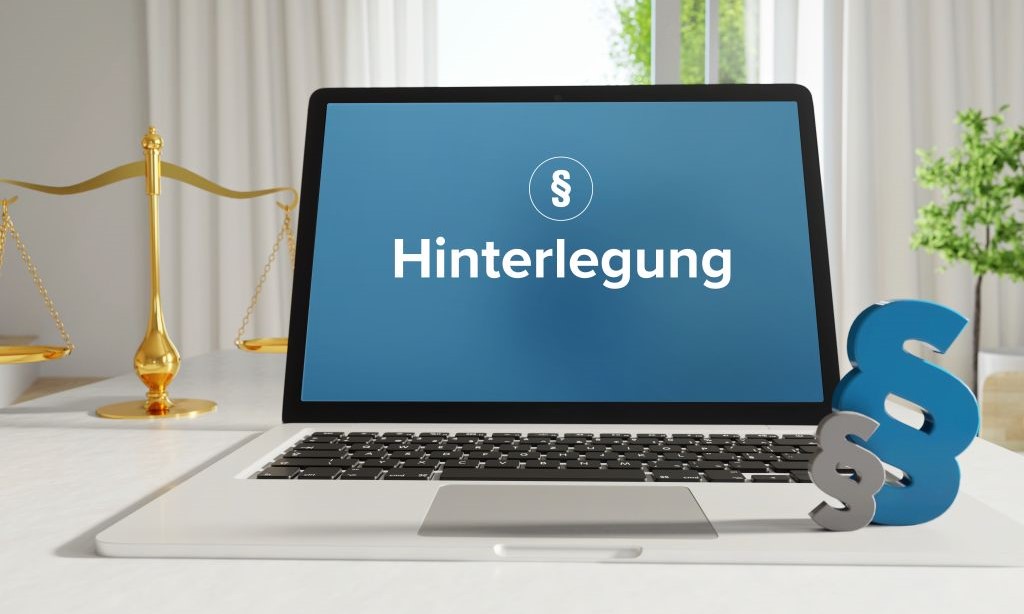 Auf einem Laptopbildschirm sieht man das Wort „Hinterlegung“ und ein Paragrafenzeichen. Neben dem Laptop stehen die Waage der Gerechtigkeit und zwei Paragrafenzeichen. 