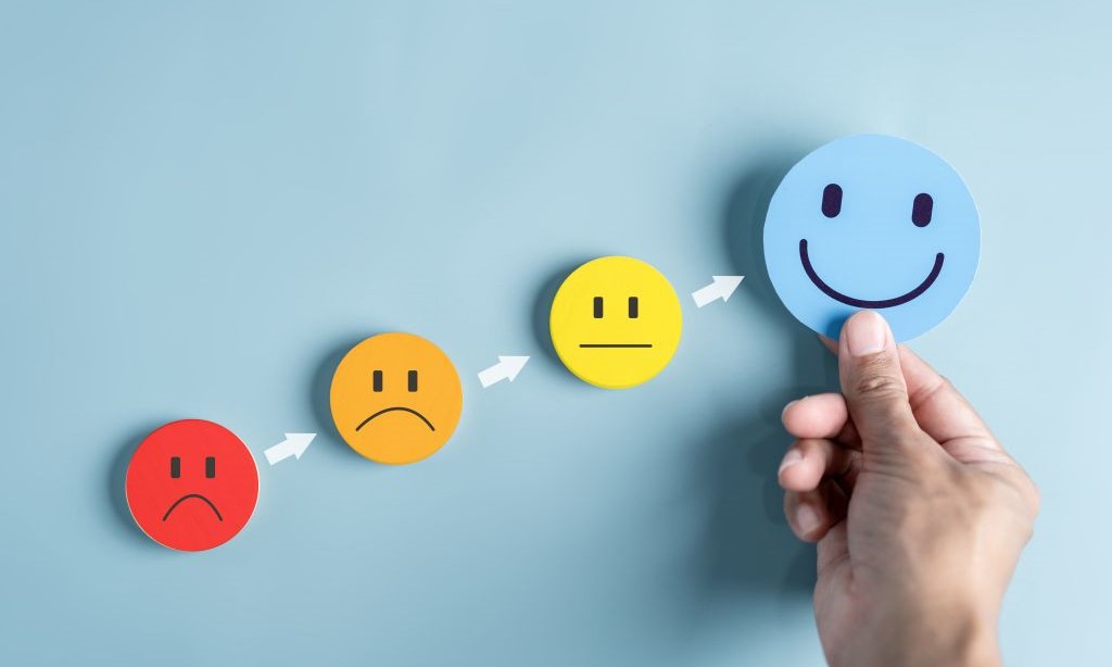Vier unterschiedliche Papier-Smileys bilden eine aufsteigende Skala vom traurigen zum lächelnden Smiley, wobei Letzteres von einer Hand hochgehalten wird.