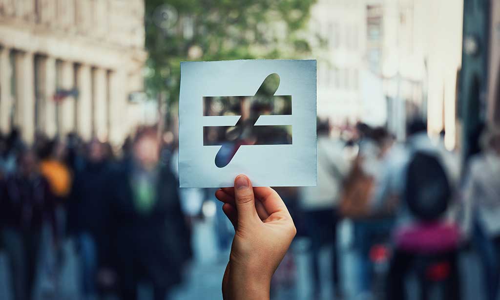 Eine Hand hält ein Papier hoch, auf dem ein Ungleich-Zeichen ausgeschnitten ist. Im Hintergrund stehen eine Gruppe Menschen.