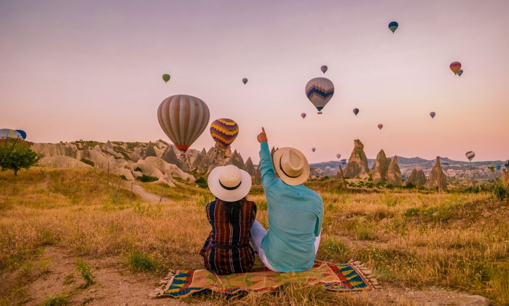 Zwei Personen sitzen in auf einer Wiese und beobachten wie Heißluftballons in der Dämmerung aufsteigen.