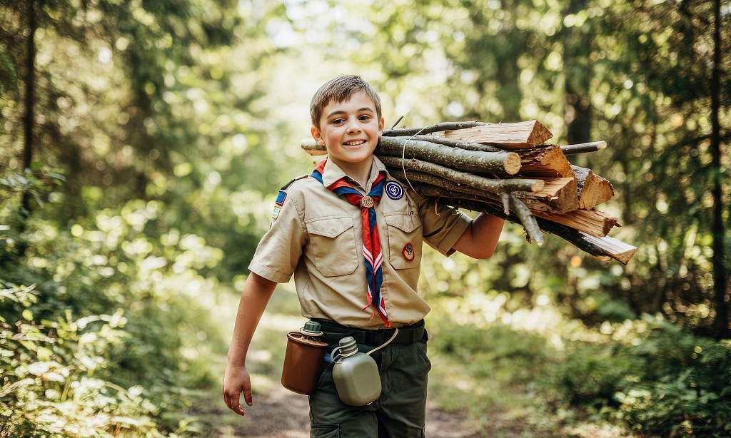 Ein Junge in Pfadfinder-Uniform trägt Holzstücke durch einen Wald. 