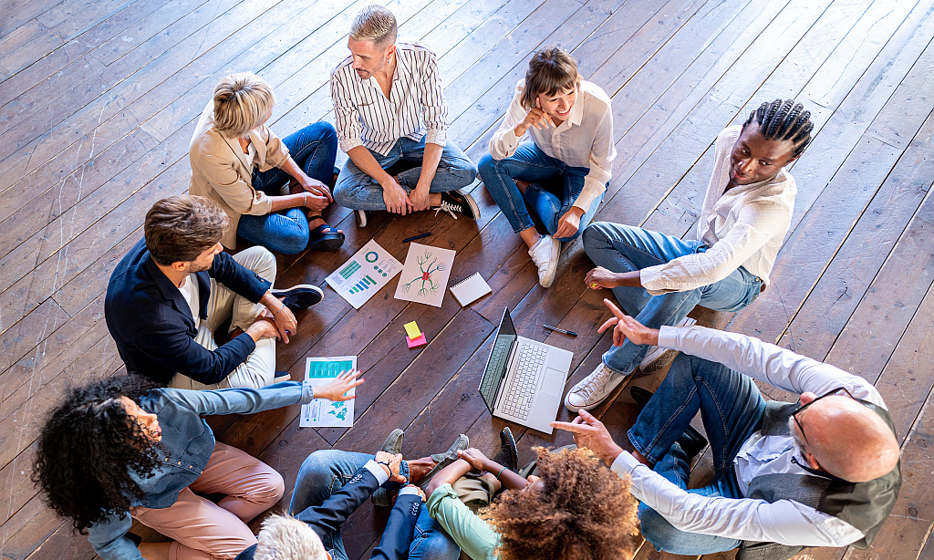 Eine Gruppe junger Menschen sitzt im Kreis am Boden und besprechen sich. In der Mitte liegen Papiere. 