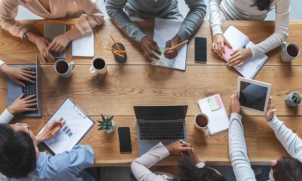 Eine Gruppe junger Menschen sitzt um einen Tisch, auf dem Smartphones, Laptops, Tassen und Schreibutensilien liegen.