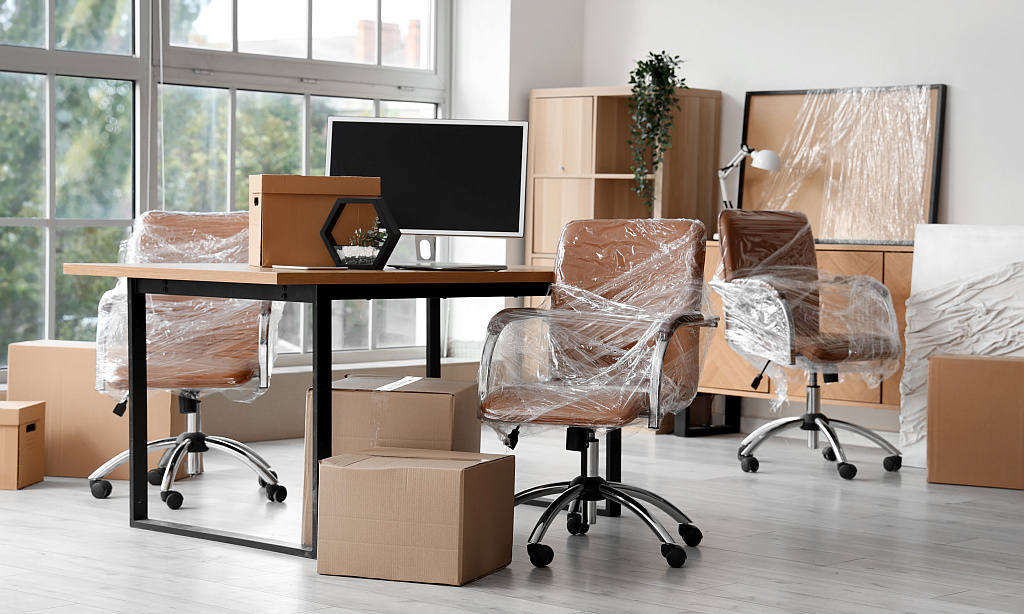 In Plastikfolie verpackte Bürostühle und Umzugskartons stehen um einen Schreibtisch herum, auf dem ein Computerbildschirm und ein Karton steht. 