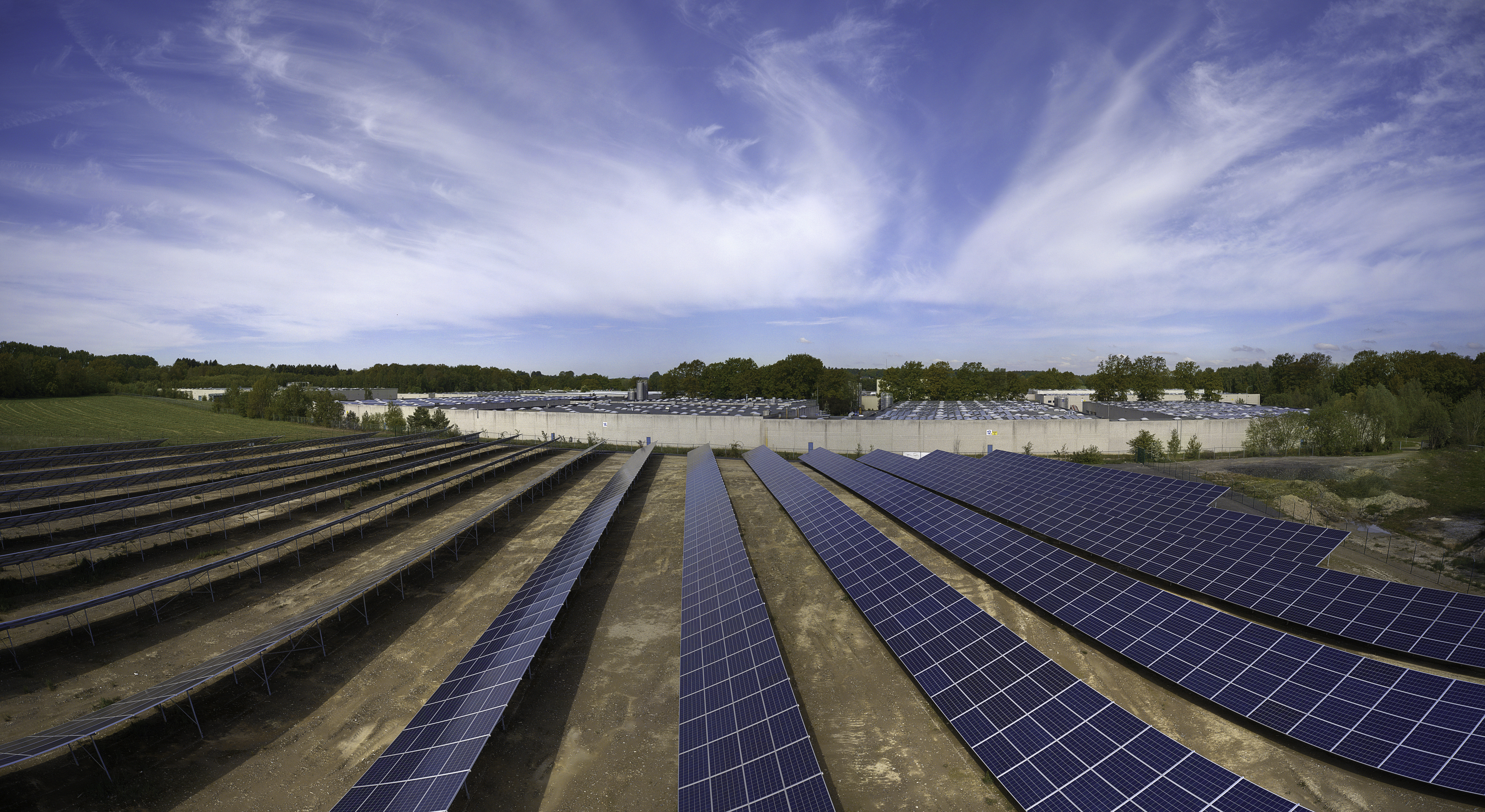 Ein Solarpark steht auf einer Wiese, im Hintergrund ist eine Fabrik zu sehen.