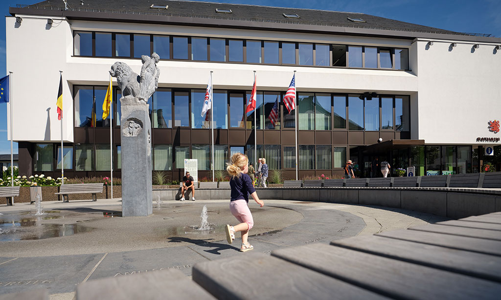 Ein Kind läuft über einen Platz mit einem Springbrunnen, im Hintergrund sieht man das Rathaus von Sankt Vith. 