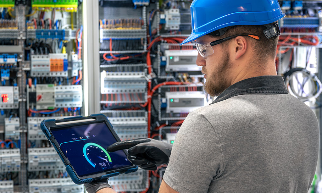Elektriker mit Tablet steht vor einem Schaltschrank. 