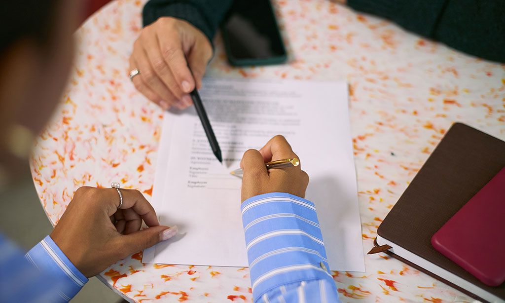 Eine Person unterschreibt ein Dokument, auf das eine weitere Person mit einem Stift in der Hand zeigt. 