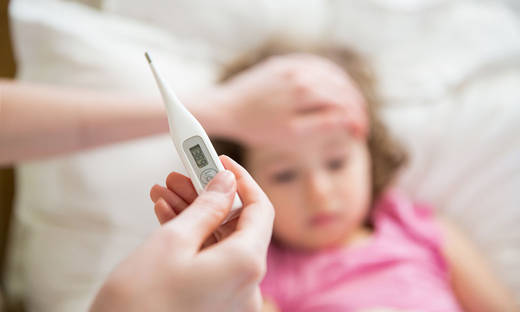 Ein Kind liegt krank im Bett, während eine Person ihm die Hand auf die Stirn legt und ein Thermometer hält.