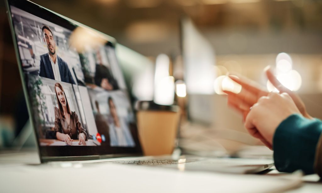 Ein aufgeklappter Laptop, auf dessen Bildschirm man eine Videokonferenz mit vier Personen sieht, steht auf einem Tisch, an dem eine Person sitzt. 