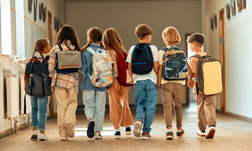 Kinder mit Schultaschen auf dem Rücken gehen in einer Reihe nebeneinander durch einen Schulflur.