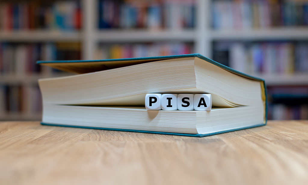 Auf einem Tisch liegt ein Buch und zwischen den Seiten liegen Holzwürfel mit Buchstaben nebeneinander und bilden das Wort „PISA“. 