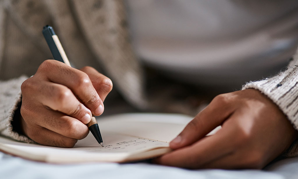 Eine Hand hält einen Stift und schreibt in ein Notizbuch, das vor der Person auf dem Tisch liegt.